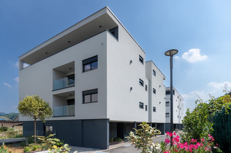 Modern multi-story apartment building with clean, minimalist design. The building has balconies on some floors and large windows. There are landscaped gardens and greenery around the building.