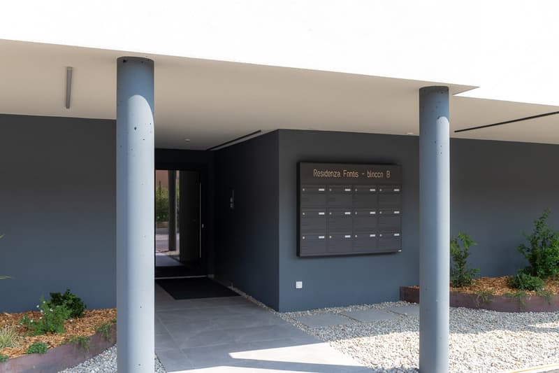 The image shows the entrance to an apartment building called 'Residenza Fontis - blocco B'. The entrance has a modern, minimalist design with gray walls, a black door, and a mailbox panel on the wall. There is a gravel path leading to the entrance and som