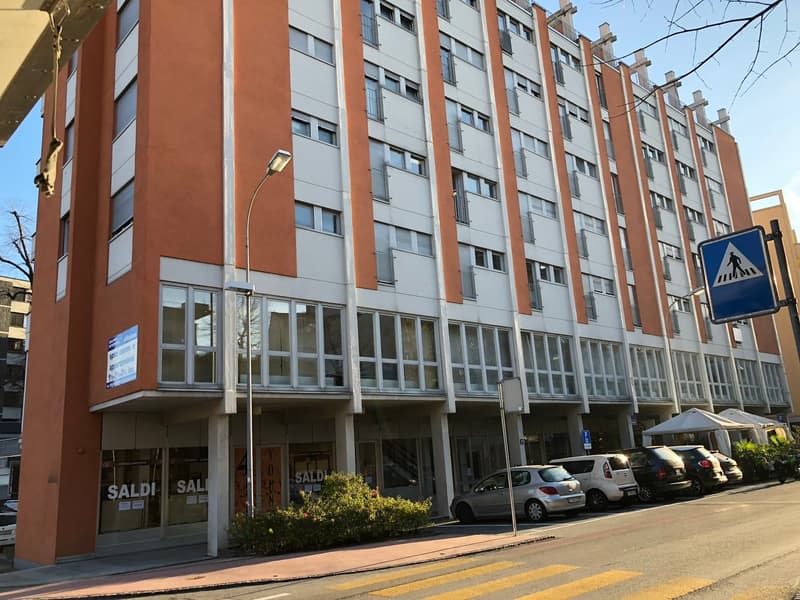 Multi-story residential building, with orange and white wall, multiple balconies, glass windows, cars parked in front