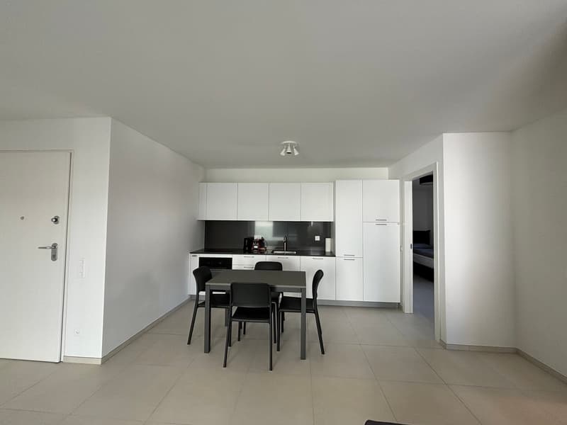 Open-concept kitchen and dining area with white cabinets, black countertops, and a dining table with chairs. Tile flooring throughout the space. Entrance door visible on the left side of the image.