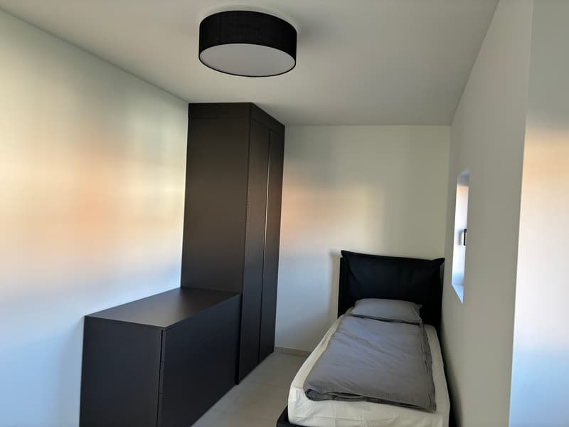 The image shows a minimalist bedroom with a black wardrobe, a black headboard, and a black ceiling light fixture. The room has white walls and a light-colored floor.