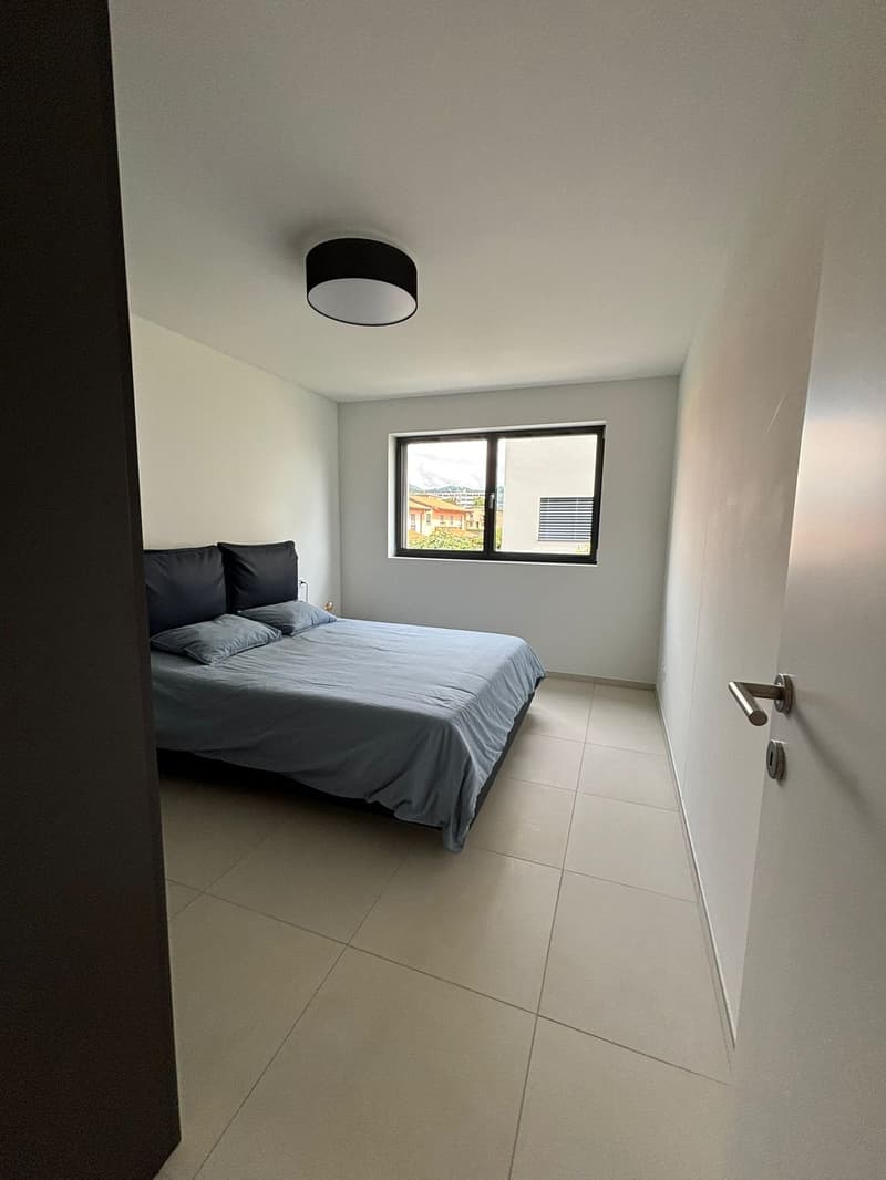 Double bed with gray comforter, two gray pillows, black headboard, large window, white walls, white tile floor, black ceiling lamp