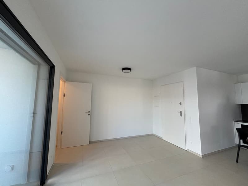 The image shows a minimalist, open-plan room with white walls, a tiled floor, and a ceiling light fixture. The room has a main entrance door and another door leading to another area. The space appears to be a living room or studio apartment with a simple,