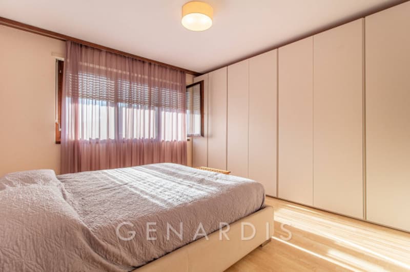 Bedroom with bed, wooden floor, white walls, large window with curtains, built-in wardrobes, ceiling lamp