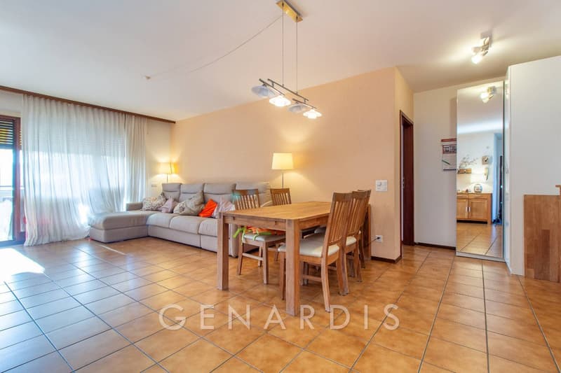 Dining room with wooden table and chairs, beige sofa, lamps, tiled floor, large window with white curtains, wooden cabinet, mirror