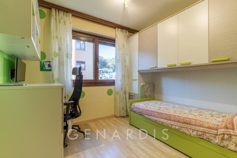 A room with a desk, chair, bed, and cabinets. The walls are painted green with white polka dots. The bed has a floral cover, and the room has a window with curtains. The floor is wooden.