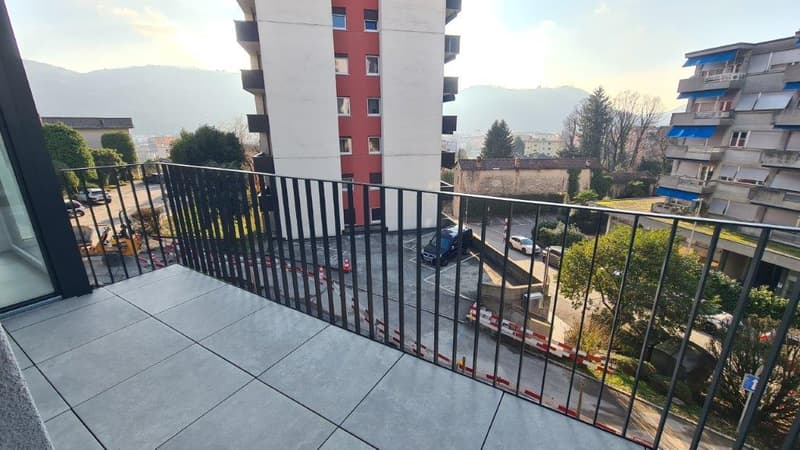 Balcony with metal railing, overlooking residential buildings and trees, with mountains in the background