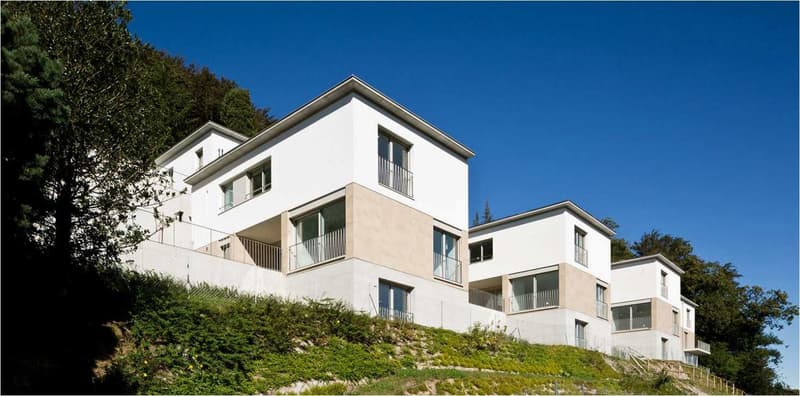 Multi-story apartment building with white exterior, balconies, and set on a hillside with trees and vegetation surrounding it.