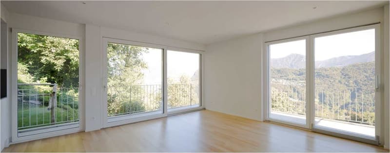 Spacious living room with large windows offering a scenic view of the surrounding greenery. The room has hardwood floors and a balcony or terrace accessible through sliding glass doors.