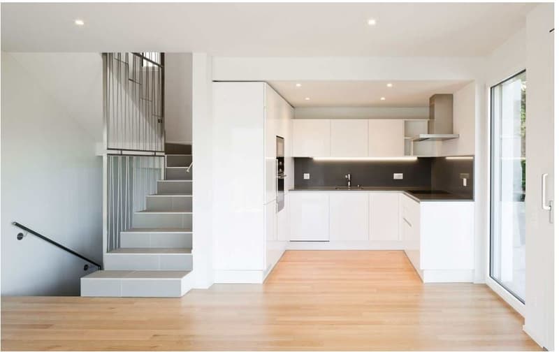 Spacious open-concept living area with white walls, hardwood floors, modern kitchen with white cabinets and a black backsplash, and a staircase with a metal railing leading to an upper level.