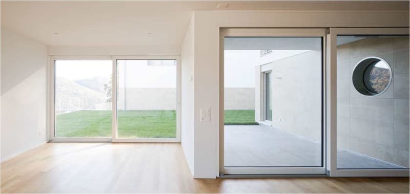 Spacious living room with large windows overlooking a grassy outdoor area, hardwood floors, and a sliding glass door leading to a balcony or terrace with a view.