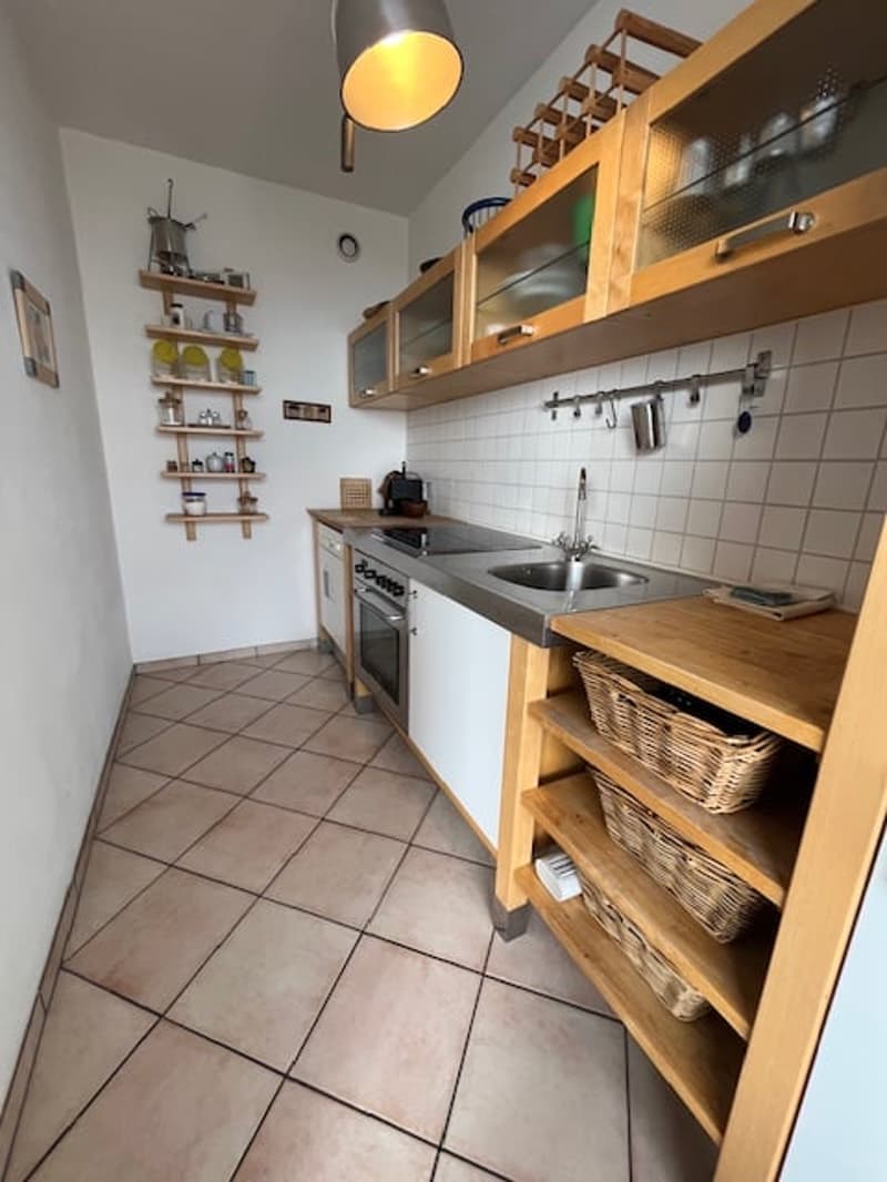 a small kitchen with tile flooring, wooden cabinets, tiled countertops, stainless steel sink and oven, and hanging shelves