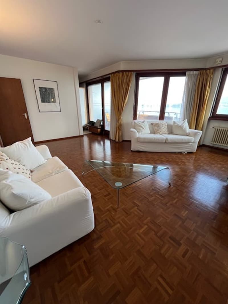 Spacious living room with white couches, glass coffee table, wooden floors, large windows, balcony with curtains, picture frame, radiator