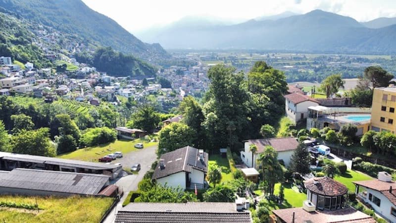 View of a mountain village with many houses and trees, several cars parked on the road, distant mountains