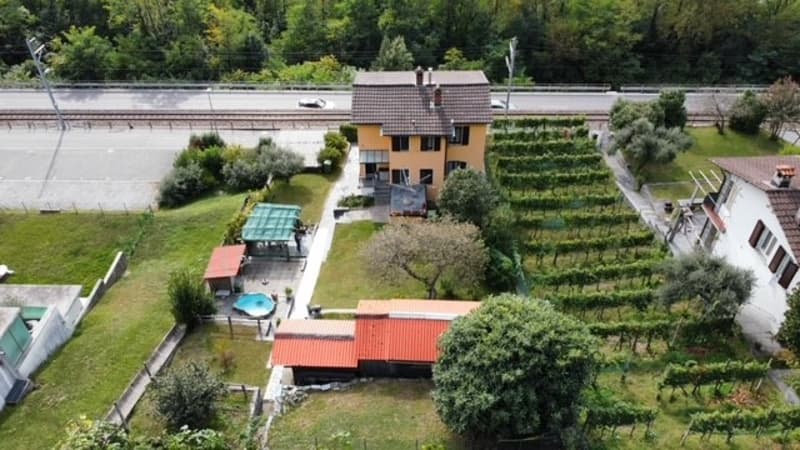 2-story house with orange roof tiles, surrounded by a garden with a swimming pool, parking space, and a balcony or terrace. The property also has a vineyard or garden area on the slope behind the house.