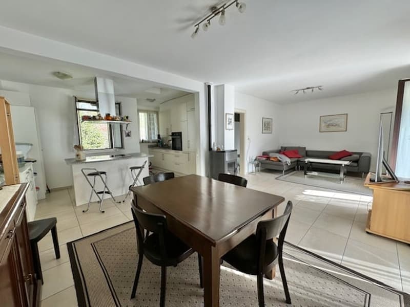 The image shows an open concept living space with a kitchen, dining area, and living room. The kitchen features modern appliances including a dishwasher. The dining table is situated in the center of the room, with a living area featuring a sofa and other