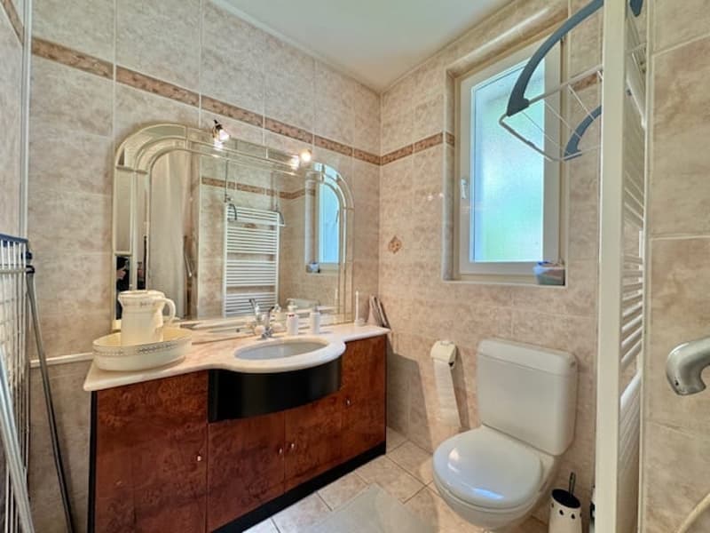 The image shows a spacious bathroom with beige tile walls, a vanity with a sink and mirror, and a toilet. The bathroom appears to be well-lit with a window providing natural light.