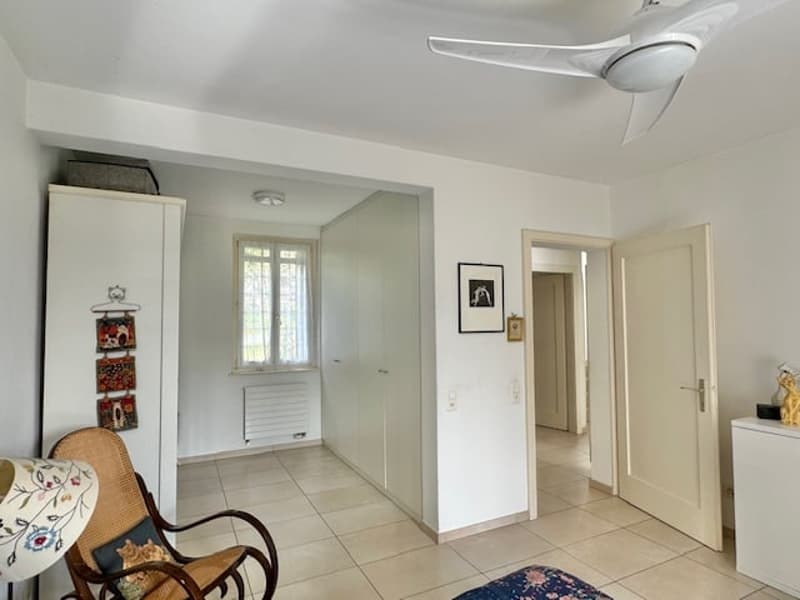 The image shows an interior of an apartment or home. The room has white walls, tile flooring, and a ceiling fan. There is a rattan chair in the foreground, and a doorway leading to another room can be seen in the background.
