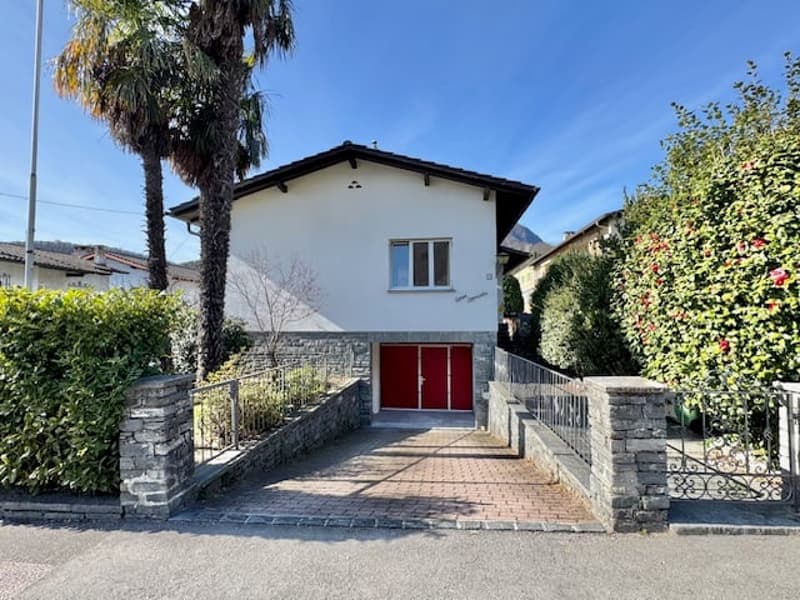 2-story white house with a red garage door, surrounded by palm trees and other vegetation. The house has a brick walkway leading to the entrance.