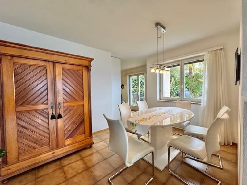 Dining area with a large wooden cabinet, a round white table, white chairs, and glass windows.