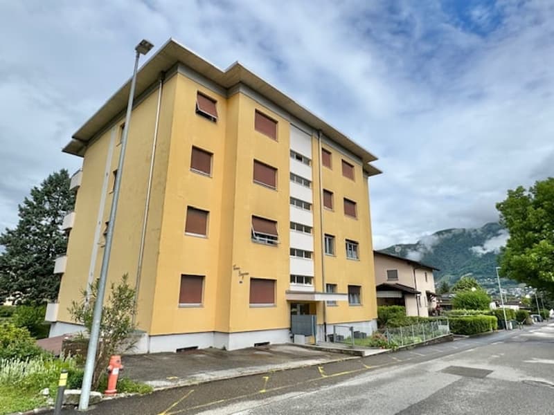 A multi-story apartment building with a yellow exterior, located on a street with trees and mountains in the background.
