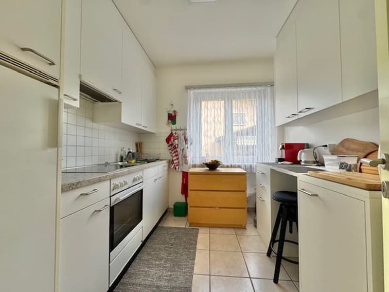 Spacious kitchen with white cabinets, tile flooring, and a dishwasher. There is a wooden cabinet island in the center and a window providing natural light.