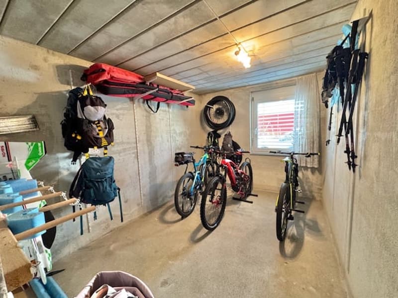 The image shows a garage or storage space with various items stored inside, including bicycles, sports equipment, and other miscellaneous items. The space appears to be cluttered but organized, with a concrete floor and a ceiling with exposed wooden beams