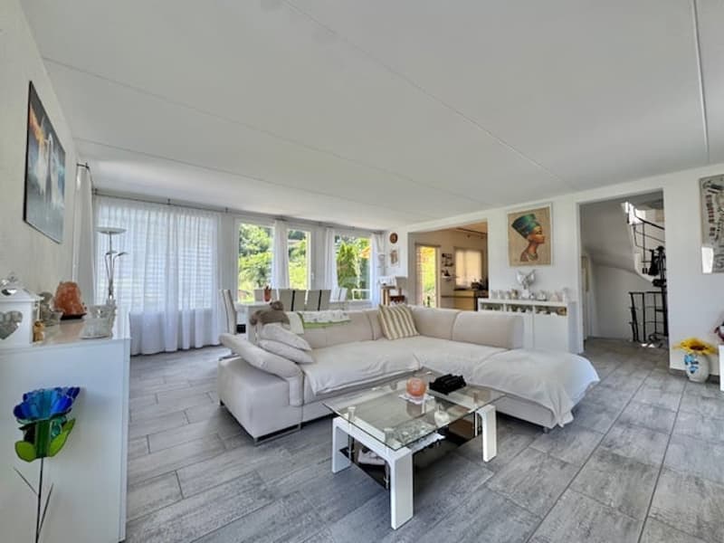 Spacious open-plan living room with large windows, white sectional sofa, glass coffee table, and tiled flooring. The room has access to a balcony or terrace area.