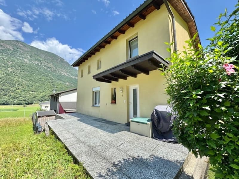 Two-story house with yellow exterior, wooden roof, balcony, and a paved driveway or parking area in front of the house. The house is surrounded by greenery and mountains in the background.
