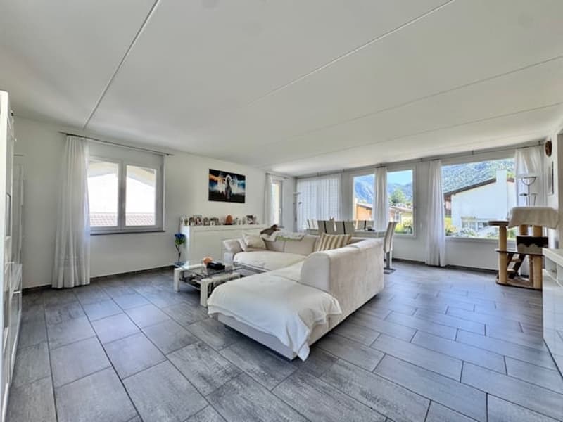 Spacious living room with large windows, white walls, hardwood-style tile flooring, white sofa, coffee table, and decorative items on shelves.