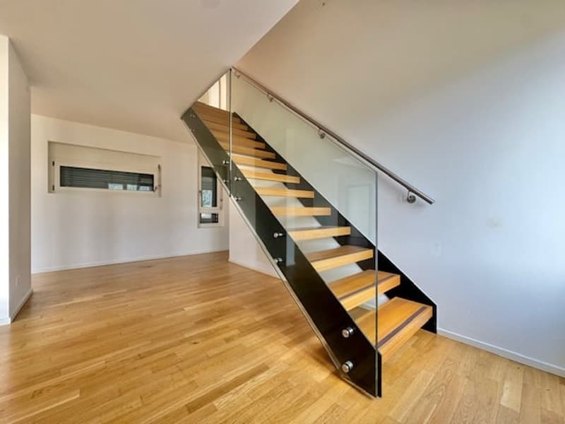 Open-concept living space with hardwood floors, glass and metal staircase leading to an upper level, and a window or door that suggests the presence of a balcony or terrace.