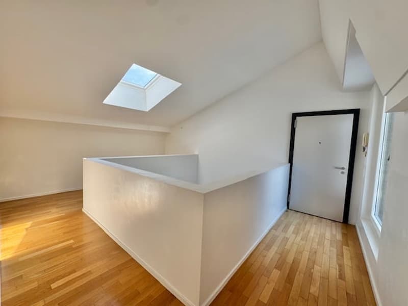 The image shows a hallway with a skylight, hardwood floors, and a white wall. There is a doorway visible at the end of the hallway.