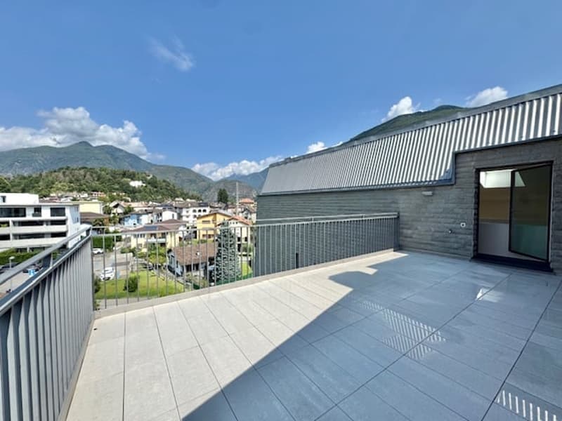 Large balcony or terrace with a view of the surrounding buildings and mountains in the background. The balcony has a concrete floor and a metal railing.