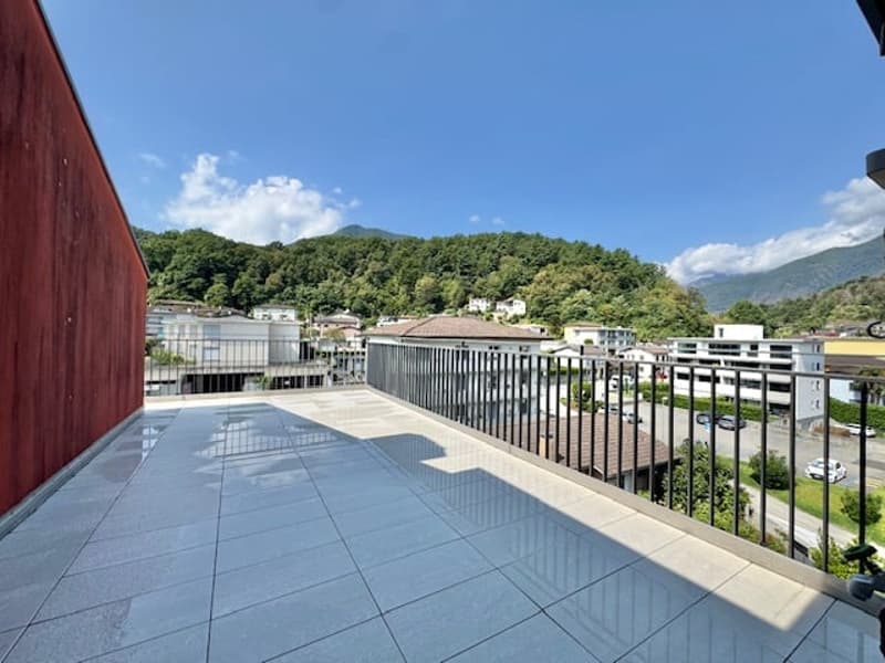 The image shows a spacious balcony or terrace with a tiled floor and metal railings. The balcony overlooks a scenic view of a town surrounded by mountains and lush greenery.