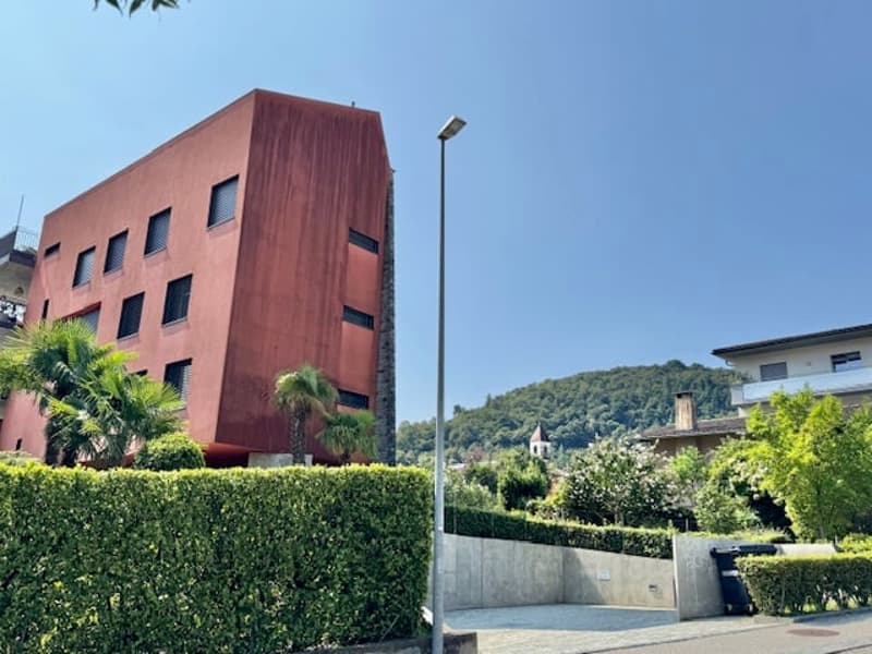 A multi-story red brick building with windows, surrounded by greenery and a clear blue sky.