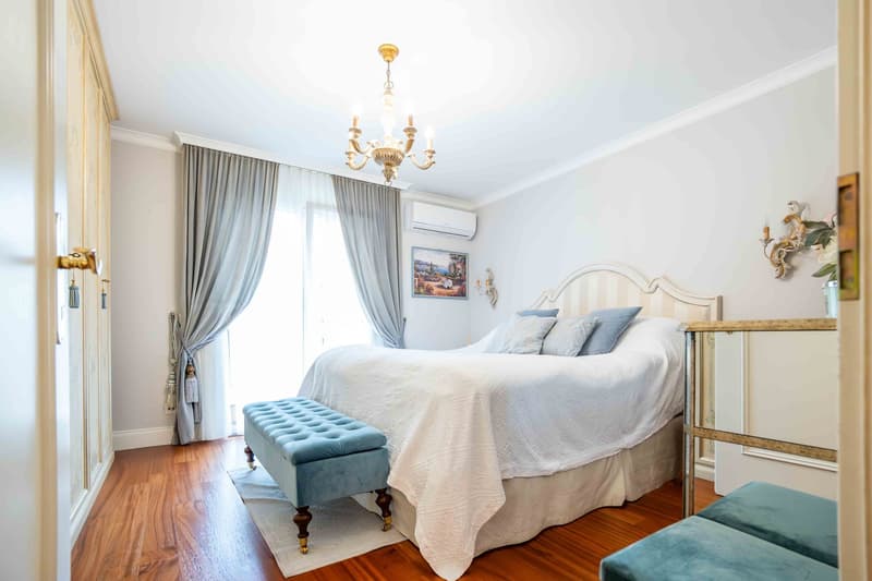 A bedroom with a large bed, white linens, chandelier, blue ottomans, wooden floor, white walls, and two windows with gray curtains