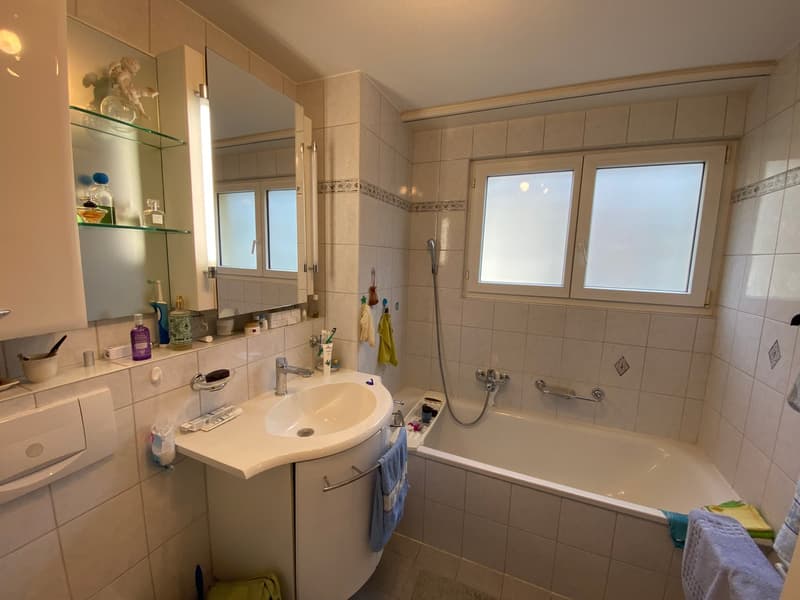 White bathroom with white tiles, a bathtub, a white sink with cabinet, mirror, glass cabinet, and shower. There are towels and a green object in the corner.