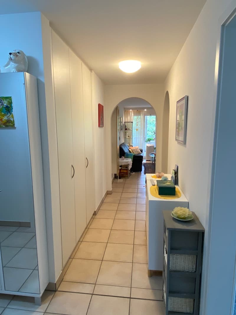 Empty hallway with white ceiling light, white tile flooring, built-in wardrobes, a cabinet with shelves, and a table with decorative items.