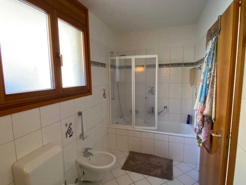 A bathroom with white tiles on the walls and floor, glass door shower, bathtub, toilet, bidet, and a window with a wooden frame.