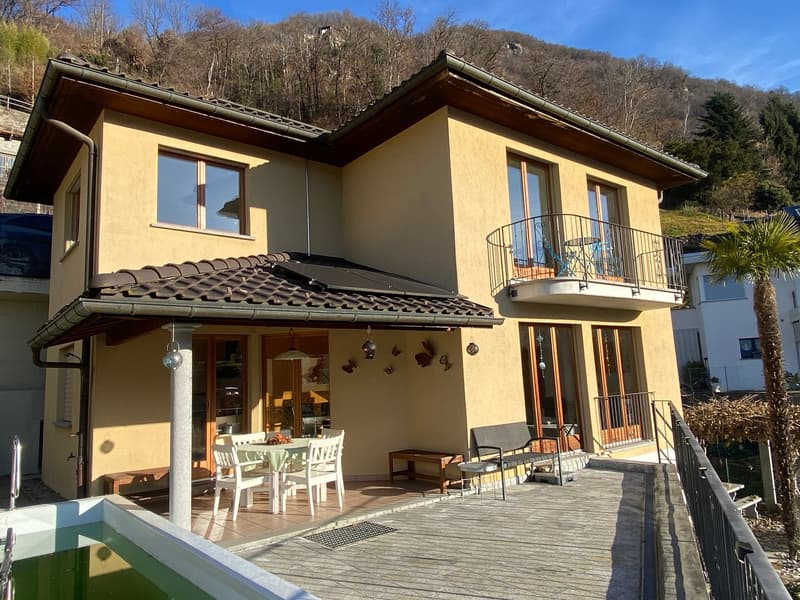 A two-story yellow house, with a brown roof, large glass windows, an entrance with awnings, a small patio with a table and chairs, a swimming pool with a handrail, a balcony with a table and chairs, a side wall with a bench and side table.