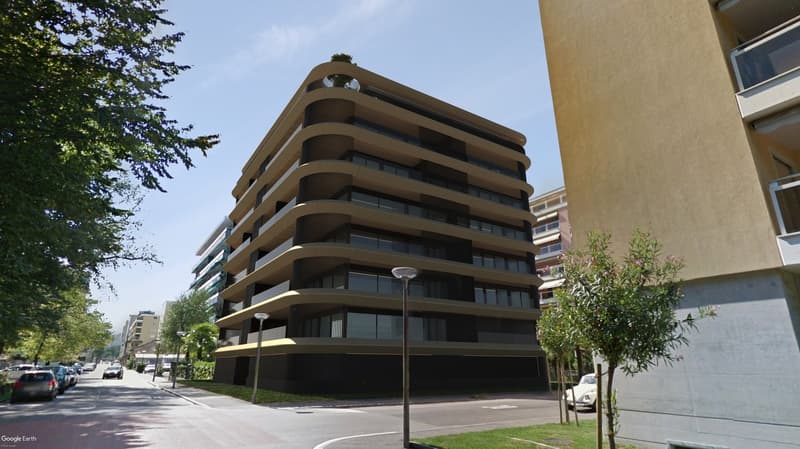 A modern multi-story apartment building with glass windows and balconies. There is a parking area and street lights on the side of the road, and trees and plants are on both sides.