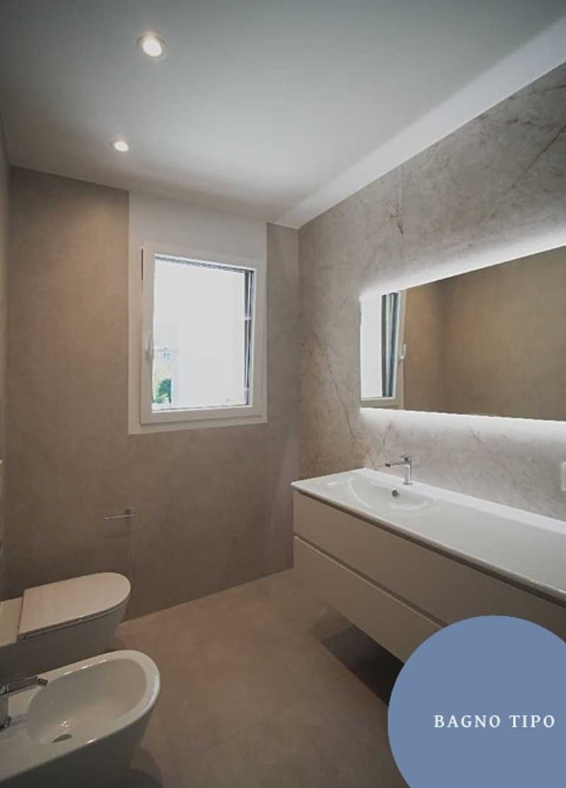A modern bathroom with white sinks, white tiles, a white toilet, and a large mirror with light fixtures