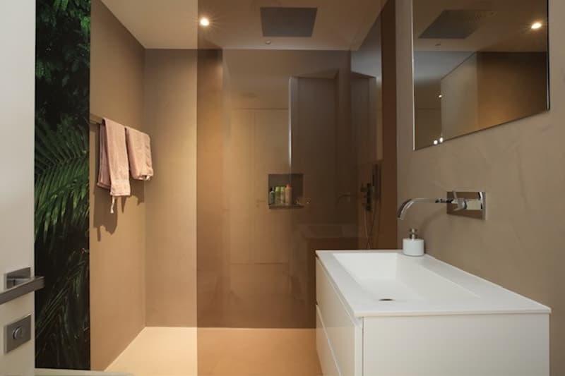 A small bathroom with a shower, a sink and a mirror, beige walls, pink towels hanging, and a decorative plant on the wall.