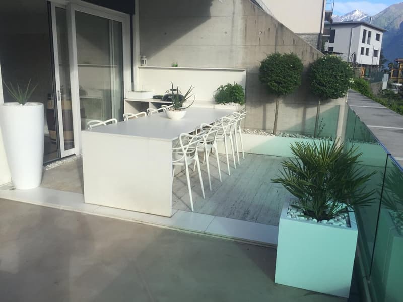 Outdoor dining area with a white table and chairs, surrounded by potted plants, a glass railing, and a view of the mountains.