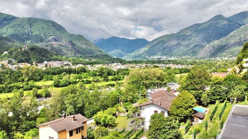 The image shows a scenic mountain landscape with a valley below. The valley is filled with lush green vegetation, including trees and other foliage. There are several buildings and houses visible in the valley, surrounded by the mountainous terrain. The s