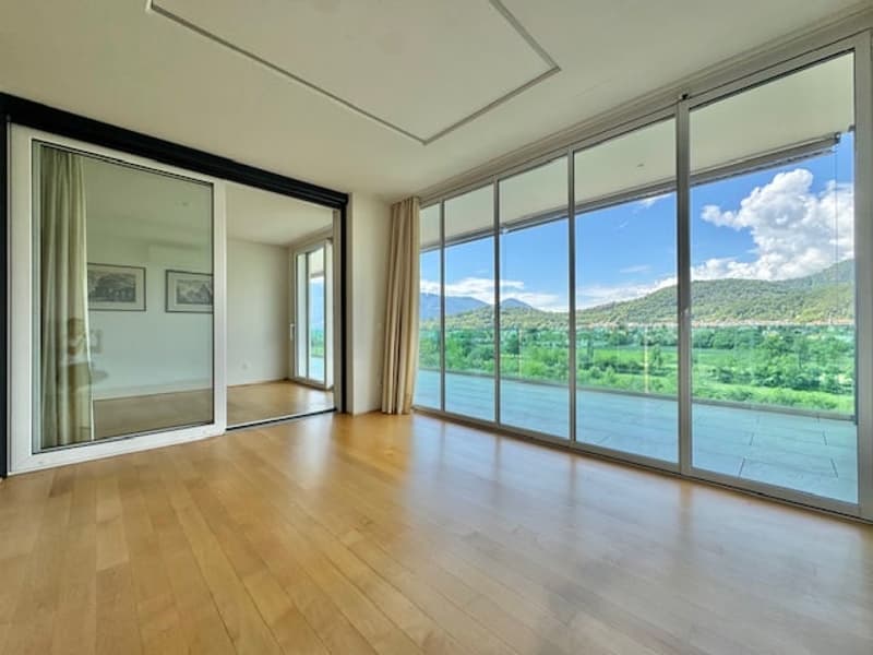 Spacious living room with large windows offering a scenic view of the surrounding mountains and greenery. The room has hardwood flooring and a sliding glass door leading to a balcony or terrace.
