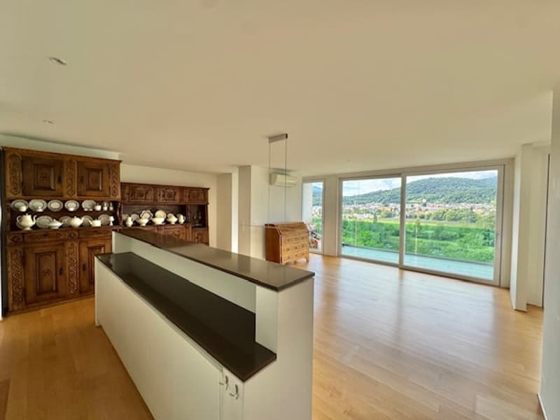 Spacious open-plan living area with large windows offering a scenic view, hardwood flooring, a built-in wooden cabinet, and a kitchen counter with a black countertop.
