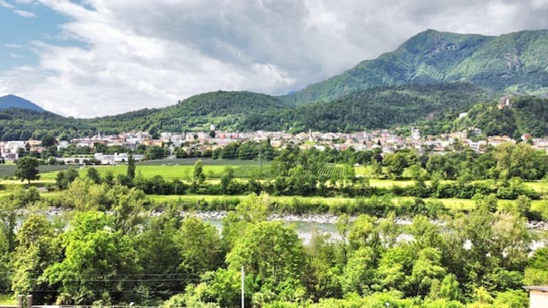 The image shows a scenic rural landscape with a small town nestled among rolling hills and mountains. The town is surrounded by lush green vegetation, including vineyards and orchards. The landscape is characterized by a mix of natural and man-made elemen