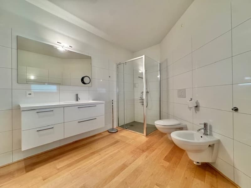 Spacious bathroom with white tile walls, wooden floor, vanity with drawers, toilet, and glass shower enclosure