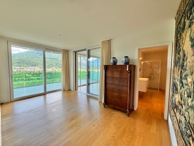 Spacious living room with large windows offering a scenic view, hardwood flooring, a wooden dresser, and an open doorway leading to another room.